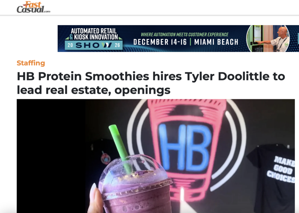 Hand holding a purple protein smoothie in a clear plastic cup with green straw and neon HB sign background